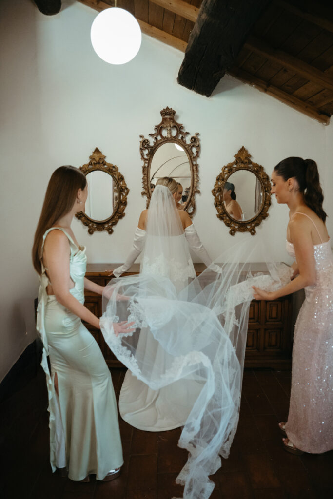 A bride smiles as two bridesmaids adjust and fluff her veil, preparing her for the ceremony.