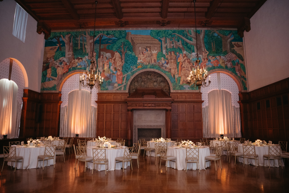 Reception hall with mural, chandeliers, and round tables.
