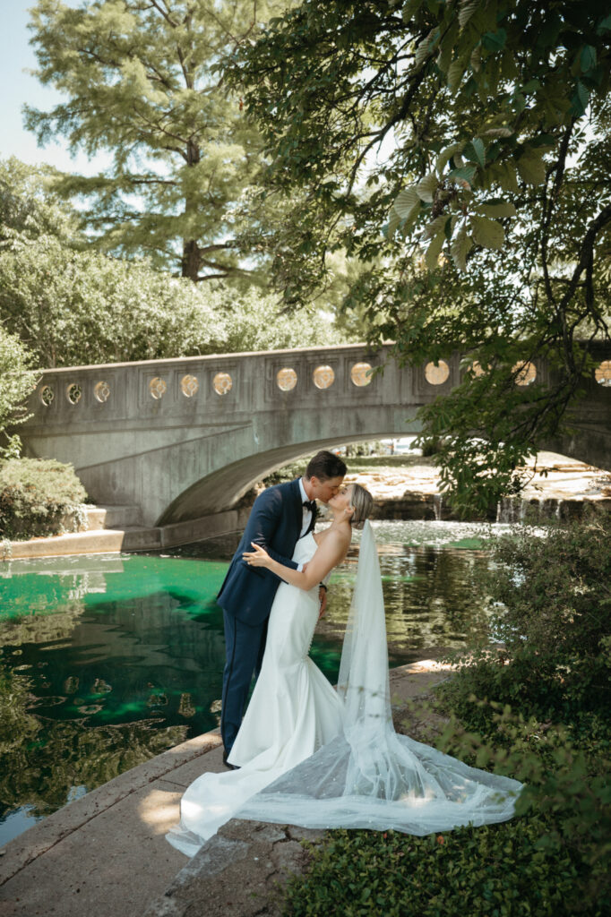 Groom dips and kisses bride near a pond.
