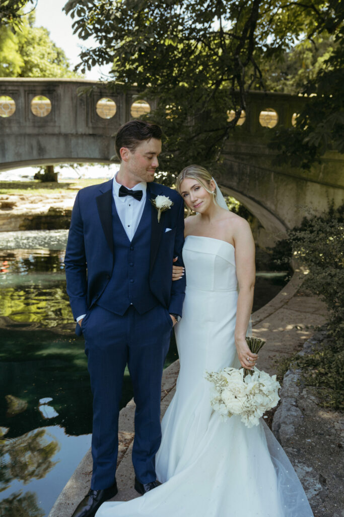 Bride leans on groom by water.
