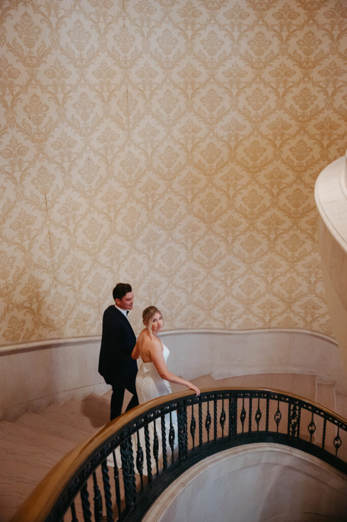 Bride and groom walking up a curved staircase with ornate wallpaper.
