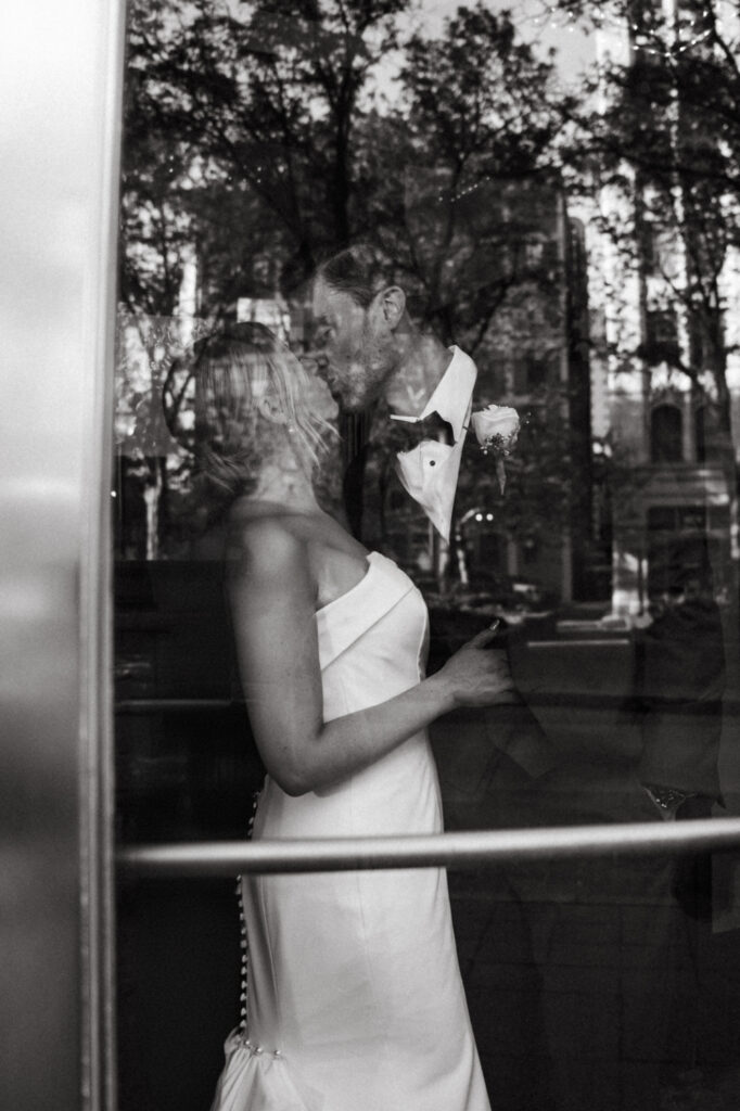 Black and white photo of couple kissing through a reflective glass door.
