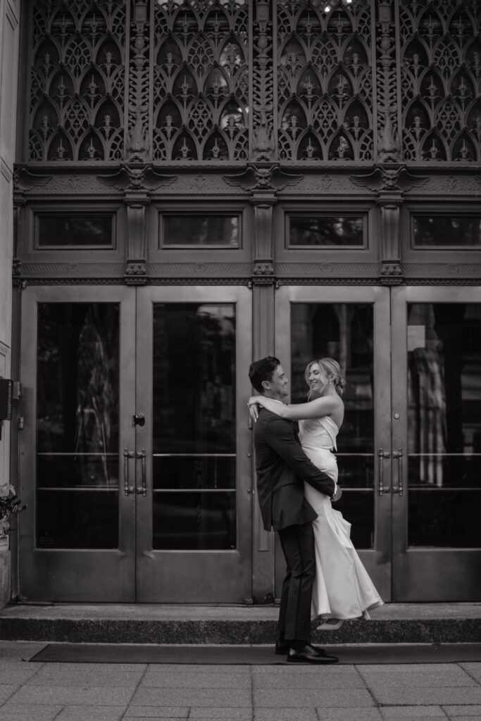 Black and white photo of groom lifting bride in front of an ornate building entrance.