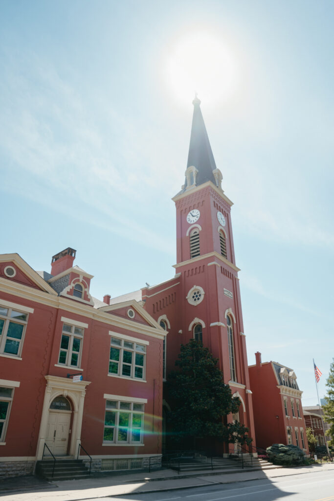 Exterior of St. Rose Catholic Church with a tall steeple, clock, and bright sun overhead.
