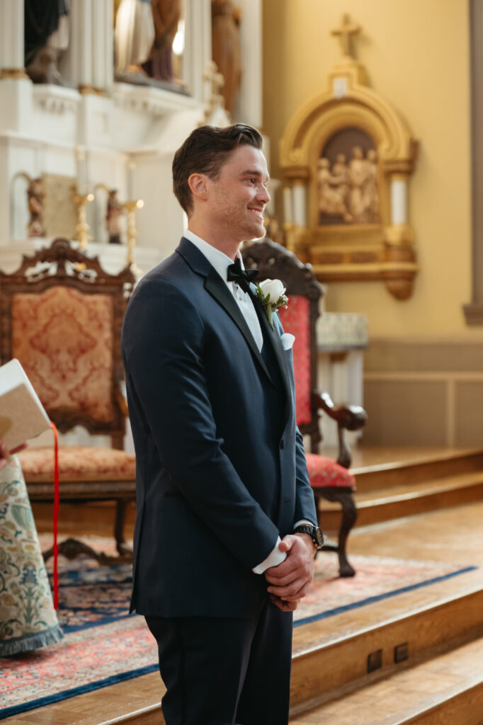 Groom stands at the altar in a navy suit, smiling as he waits for his bride at St Rose Catholic Church






