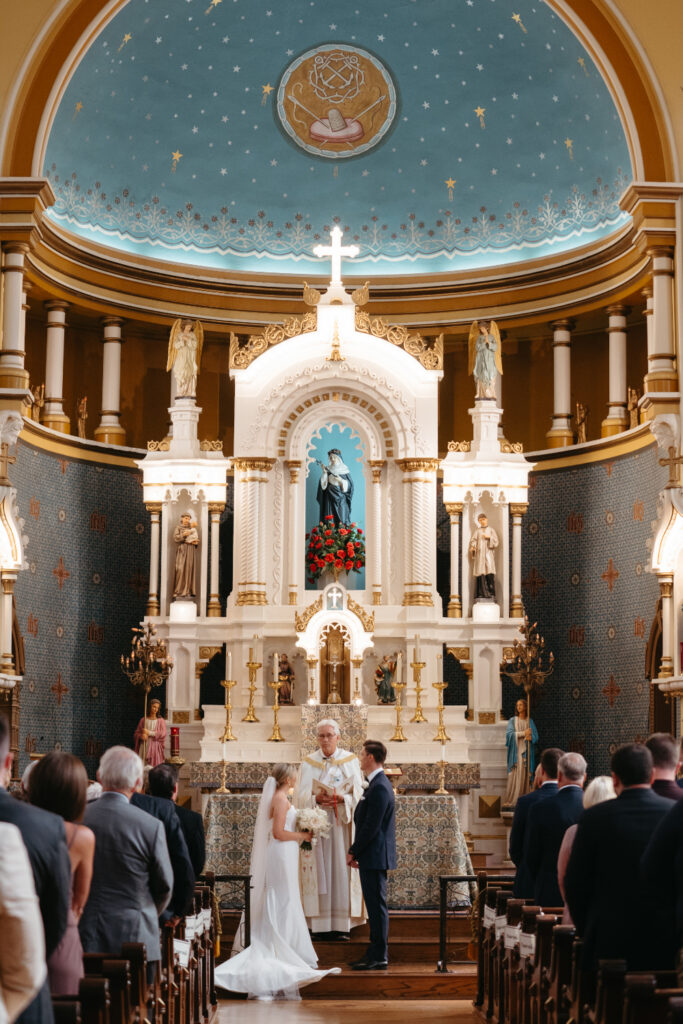 Bride and groom stand at the church altar with the priest, guests watching.
