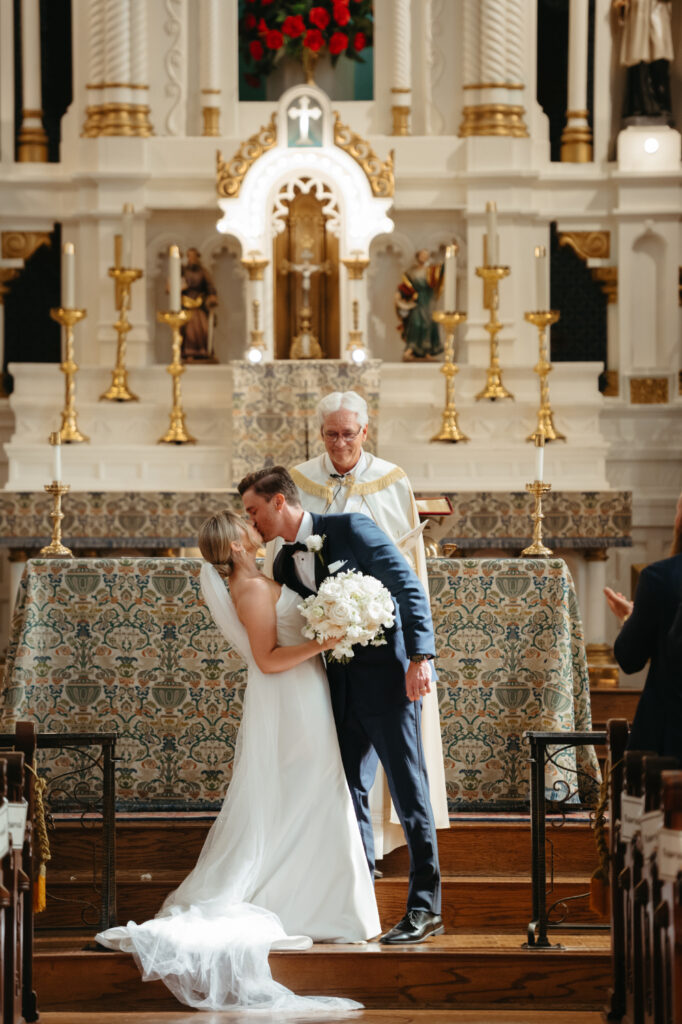 Newlyweds kiss in front of the altar, priest smiling behind them.
