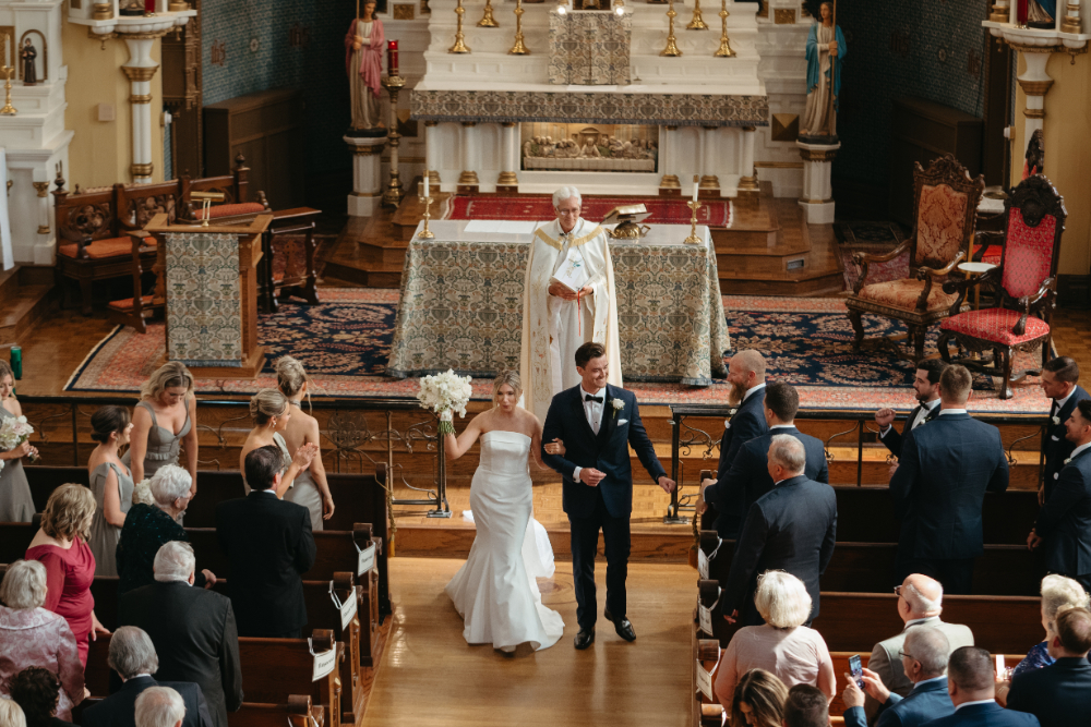 Bride and groom walk down the aisle, guests cheering.
