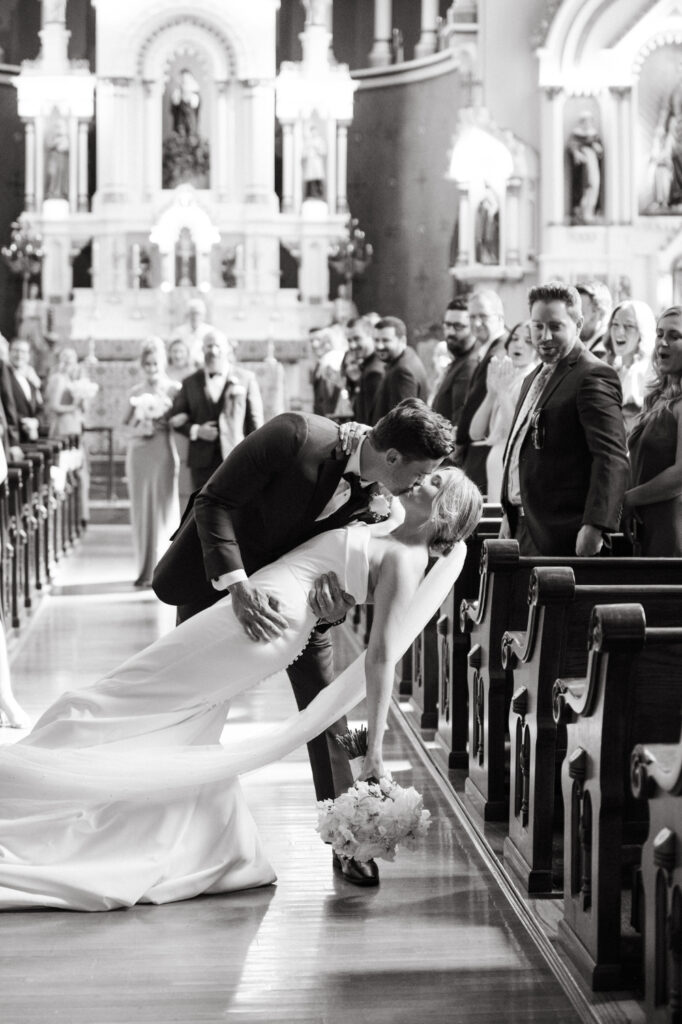 Groom dips and kisses the bride in the aisle, guests reacting.