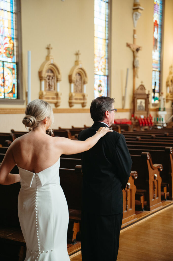 Bride places a hand on her father’s shoulder for a first look in St Rose Catholic Church.
