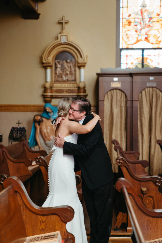 Bride and father embrace inside St Rose Catholic Church near wooden pews.
