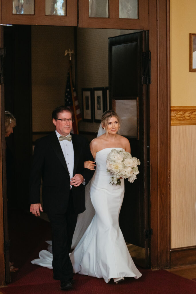 Bride and father walk arm-in-arm through church doors, bride holding white bouquet.
