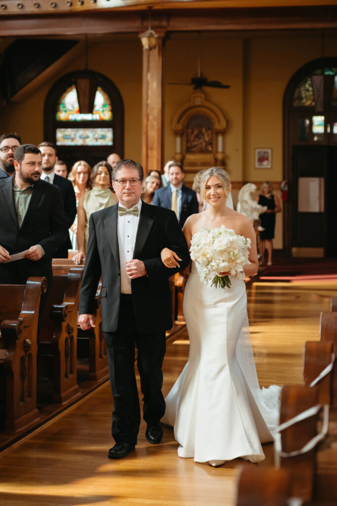 Bride and father walk down the aisle, guests watching.
