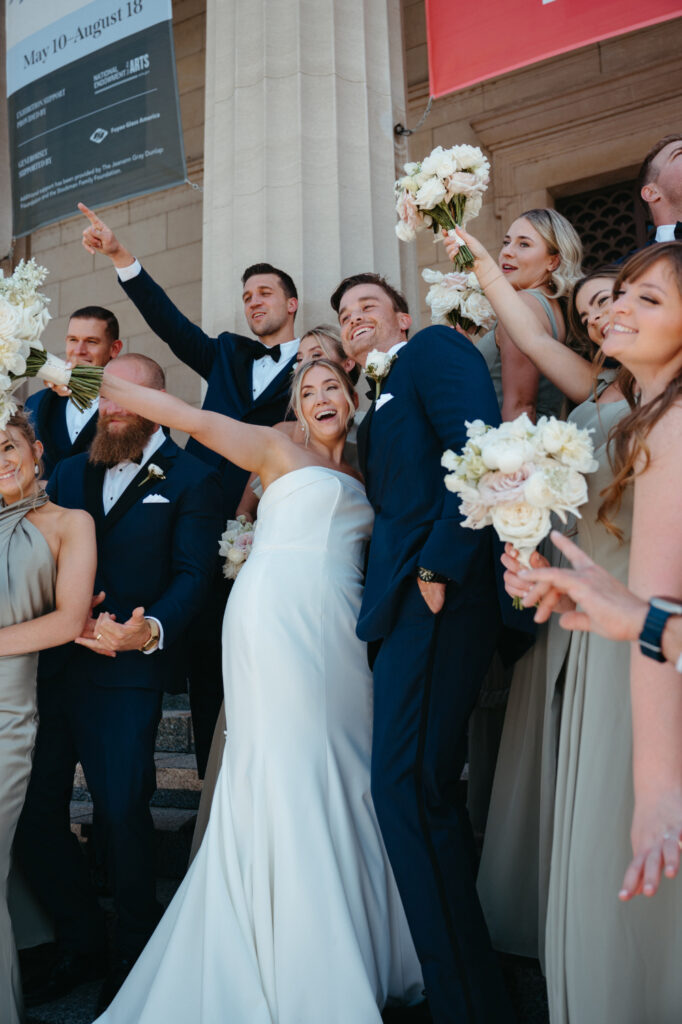 Bride and groom celebrate with their wedding party on the steps of a building, holding bouquets and smiling.
