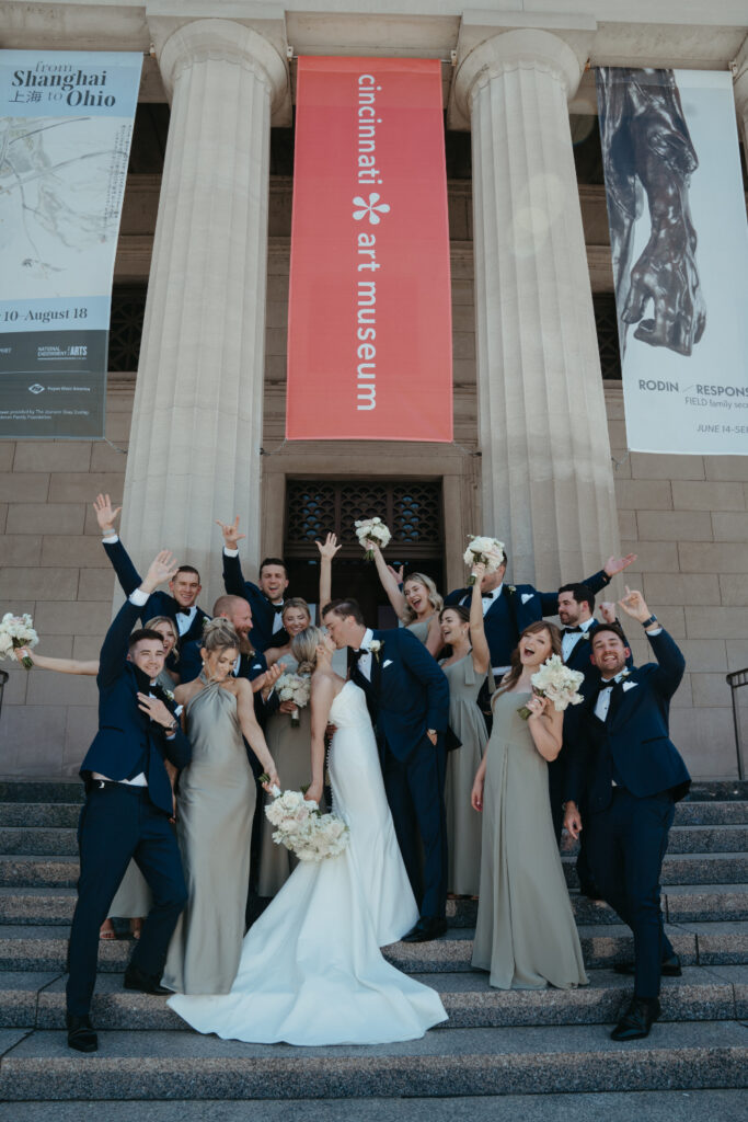 Bride and groom kiss while surrounded by cheering bridesmaids and groomsmen on the steps of the Cincinnati Art Museum, with banners hanging from the columns.