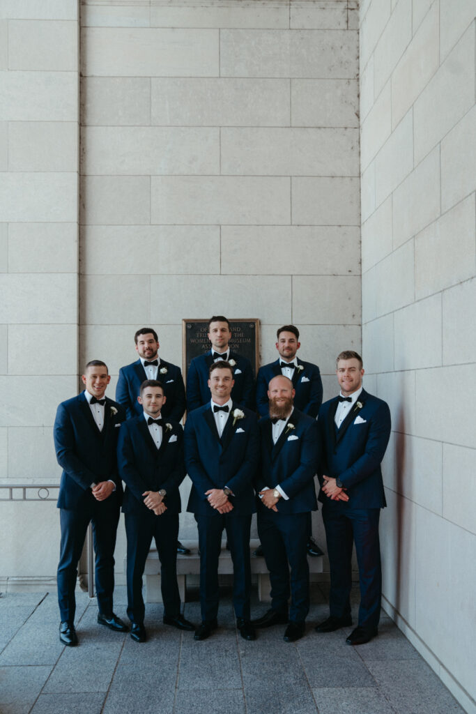 Groomsmen in navy suits posing against a stone wall.
