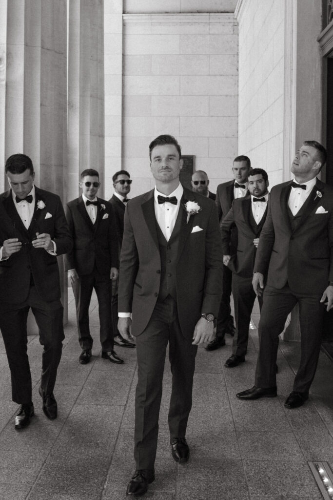 Black and white photo of groom leading groomsmen in tuxedos.
