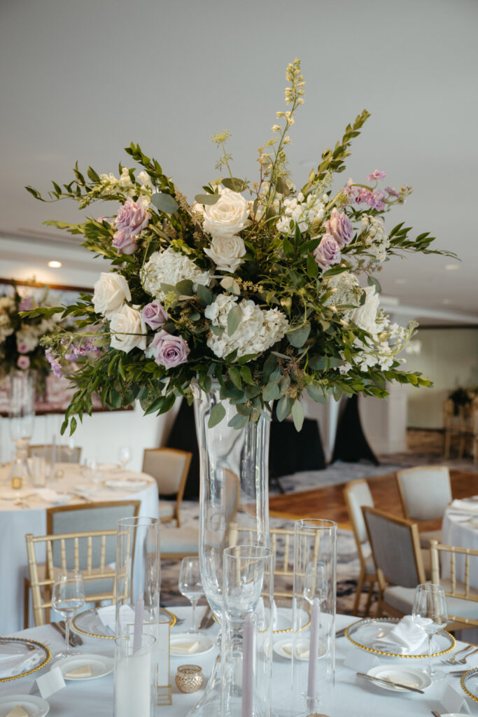 A close-up of a tall, glass vase centerpiece filled with white and lavender roses, eucalyptus, and greenery, surrounded by elegant table settings with gold accents and candlelit decor.