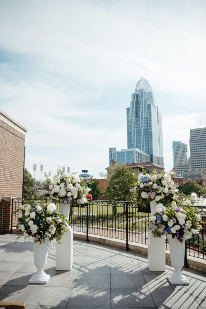 A rooftop wedding ceremony setup in Cincinnati, featuring large floral arrangements in white vases with the city skyline, including a tall glass building, in the background | best season to get married in cincinnati