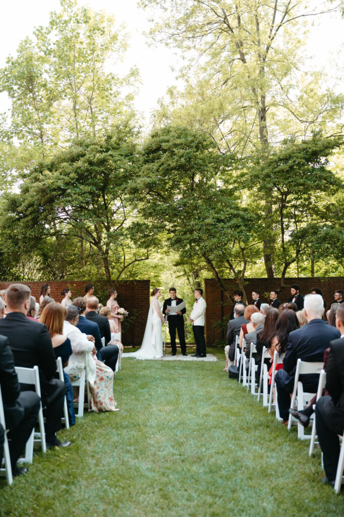 An outdoor wedding ceremony with guests seated on white chairs, facing a couple exchanging vows under a green canopy of trees and brick walls in the background.