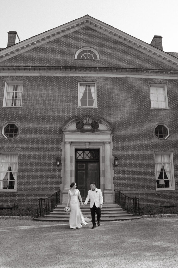 A black and white photo of a bride and groom walking hand in hand in front of a stately brick mansion with a grand entrance, large windows, and elegant architectural details.