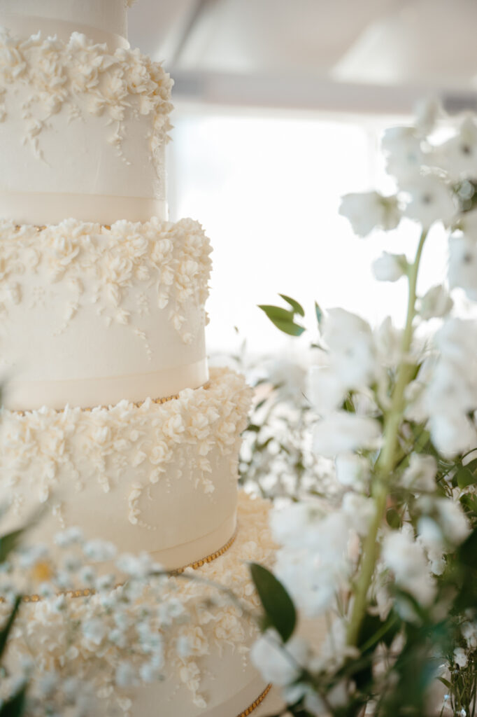 A close-up of a white, multi-tiered wedding cake with intricate floral icing details, surrounded by white flowers and greenery, softly lit by natural light.