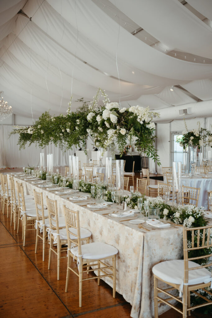 A wedding reception setup inside a tent with draped white fabric, featuring long tables with gold chairs, white and green floral arrangements, tall candles, and chandeliers in the background | the best season to get married in cincinnati