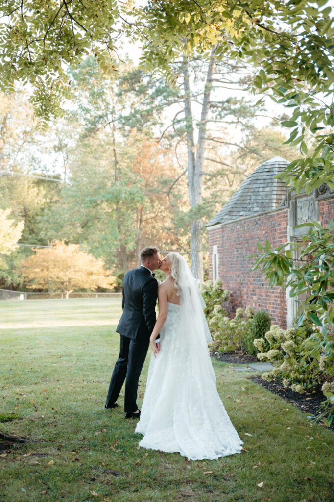 A bride and groom share a kiss in a garden with fall foliage, next to a brick building with a shingled roof. The bride wears a lace gown and veil, while the groom wears a black suit.