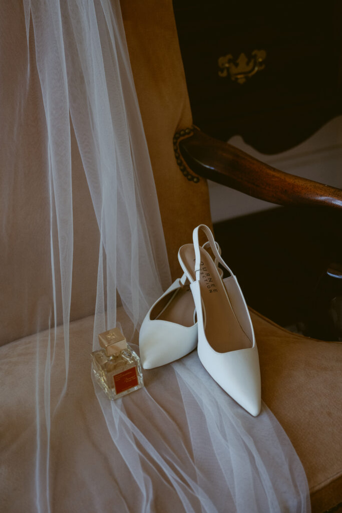 Bridal heels and perfume rest atop a vintage chair, draped with a sheer, flowing veil.