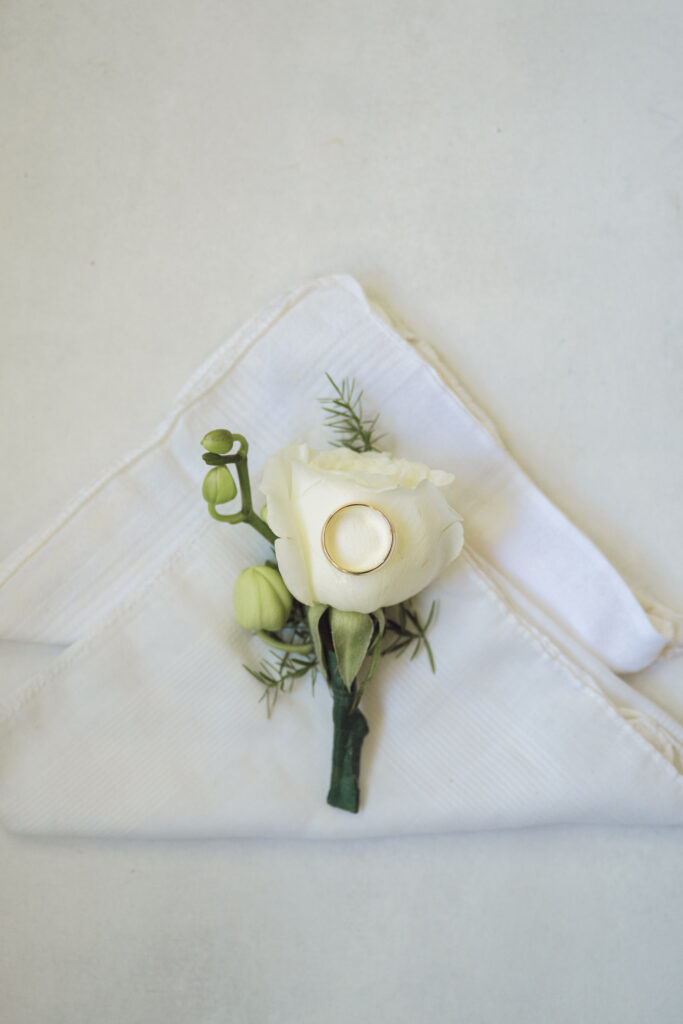 A gold wedding band rests atop a white rose boutonniere, carefully placed on a folded handkerchief.
