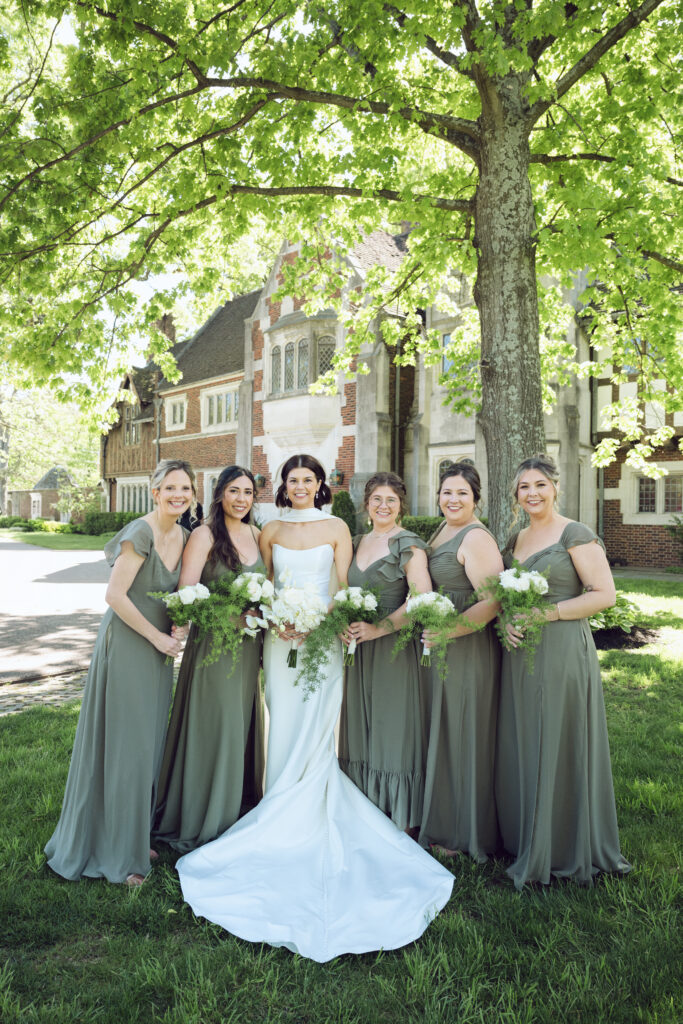 The bride stands surrounded by her bridesmaids under a leafy tree, all beaming in elegant gowns.