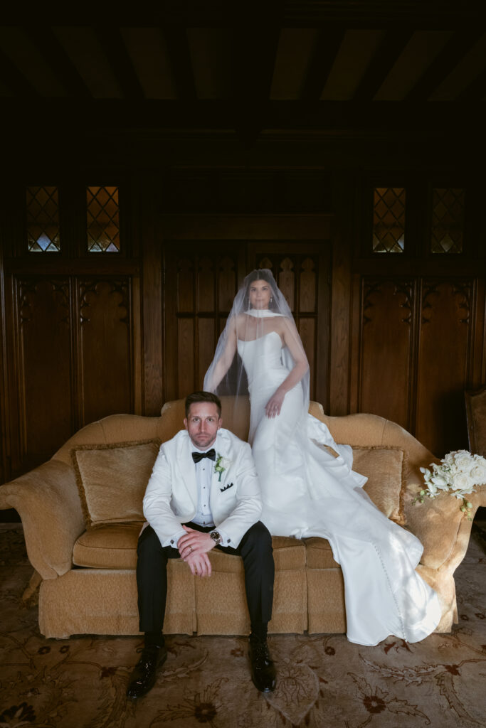 The bride stands poised in her gown and veil while the groom sits confidently on a vintage sofa.