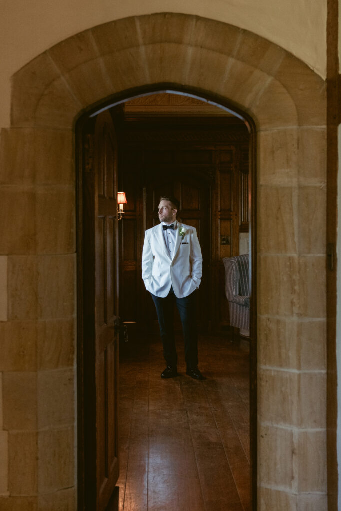 The groom stands confidently in a white tuxedo, framed by a stone archway and moody lighting.