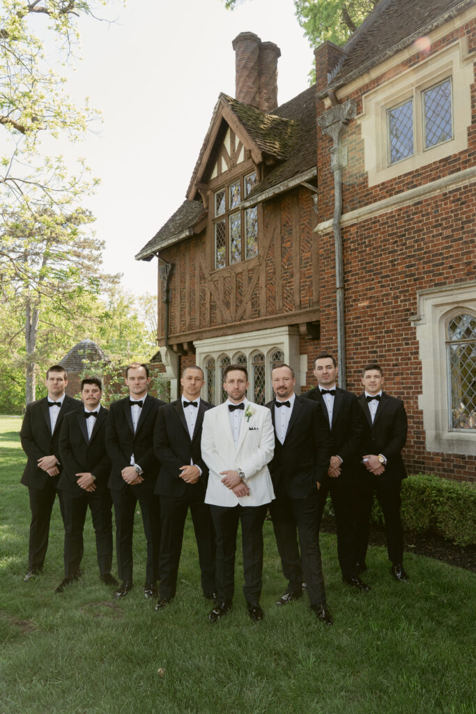 The groom stands confidently with his groomsmen in black tuxedos, posed against a historic estate backdrop.