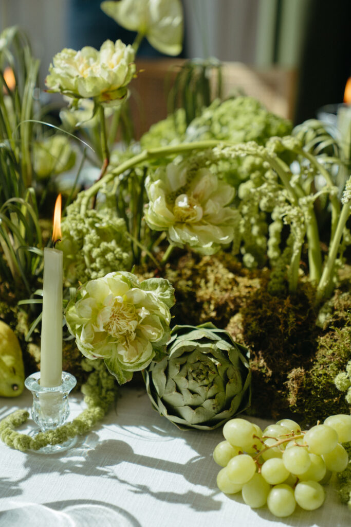 Lush green floral centerpiece with artichokes, grapes, and taper candles on a white tablecloth.