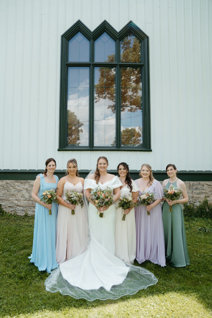 wedding-color-palette Bride and bridesmaids pose outdoors wearing pastel gowns, each holding lush bouquets in front of chapel.
