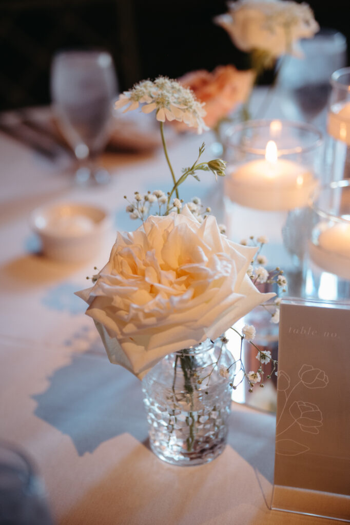 wedding-color-palette Soft white rose in clear textured vase surrounded by delicate blooms and glowing candles on table.