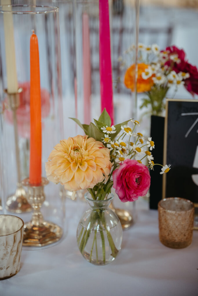 Colorful wedding table decor with bright taper candles, small glass vases of fresh flowers in pink, peach, and yellow, and gold candle holders on a white tablecloth.