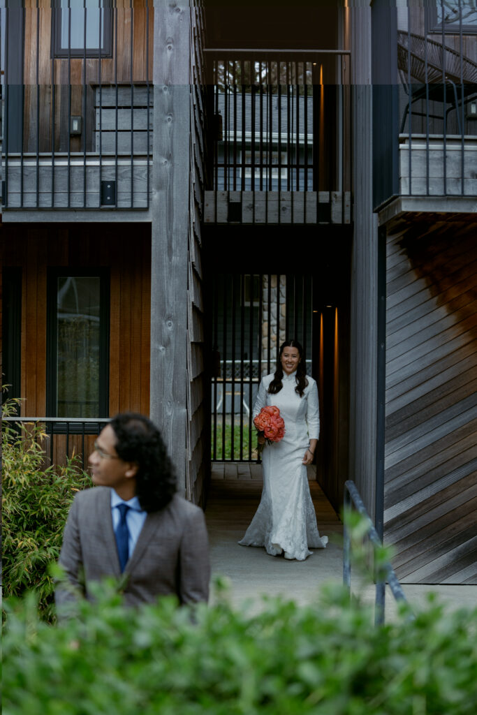 Bride walks toward groom for their first look, holding a coral bouquet and smiling with anticipation.