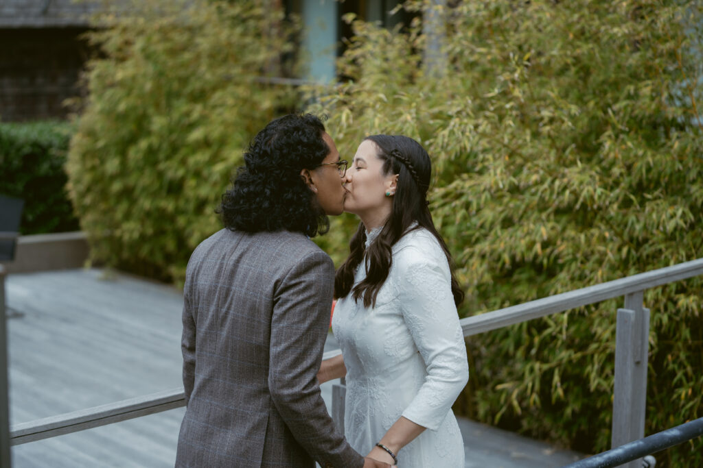 Bride and groom share a sweet kiss during their first look, surrounded by lush greenery and joy.