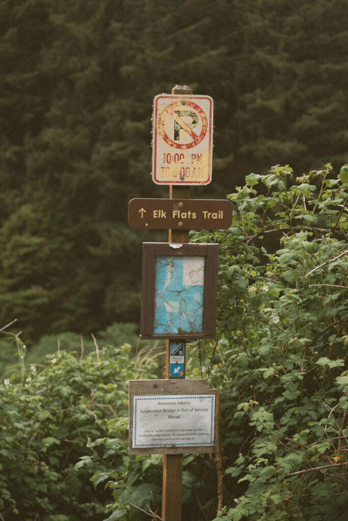 A rustic trailhead sign marks the entrance to Elk Flats Trail, surrounded by lush greenery and forest. A weathered notice alerts hikers that the suspension bridge ahead is currently out of service.