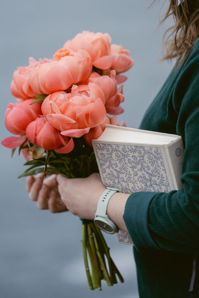 A person holds a bouquet of coral peonies and a floral-patterned book, standing near the water in a green long-sleeve shirt.