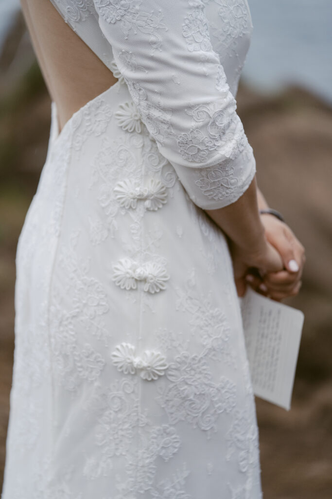 Close-up of a bride’s lace wedding dress with floral details and buttons, as she holds a small card during an outdoor ceremony.