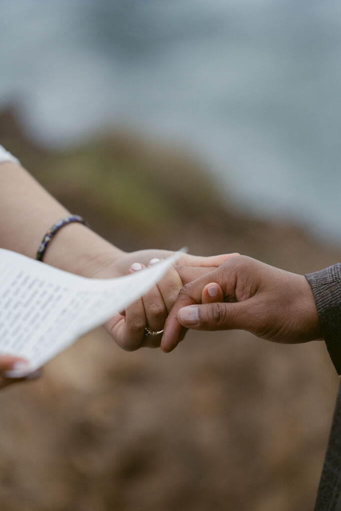 Close-up of a couple holding hands during their vows, with one partner holding a handwritten note against a blurred ocean backdrop.