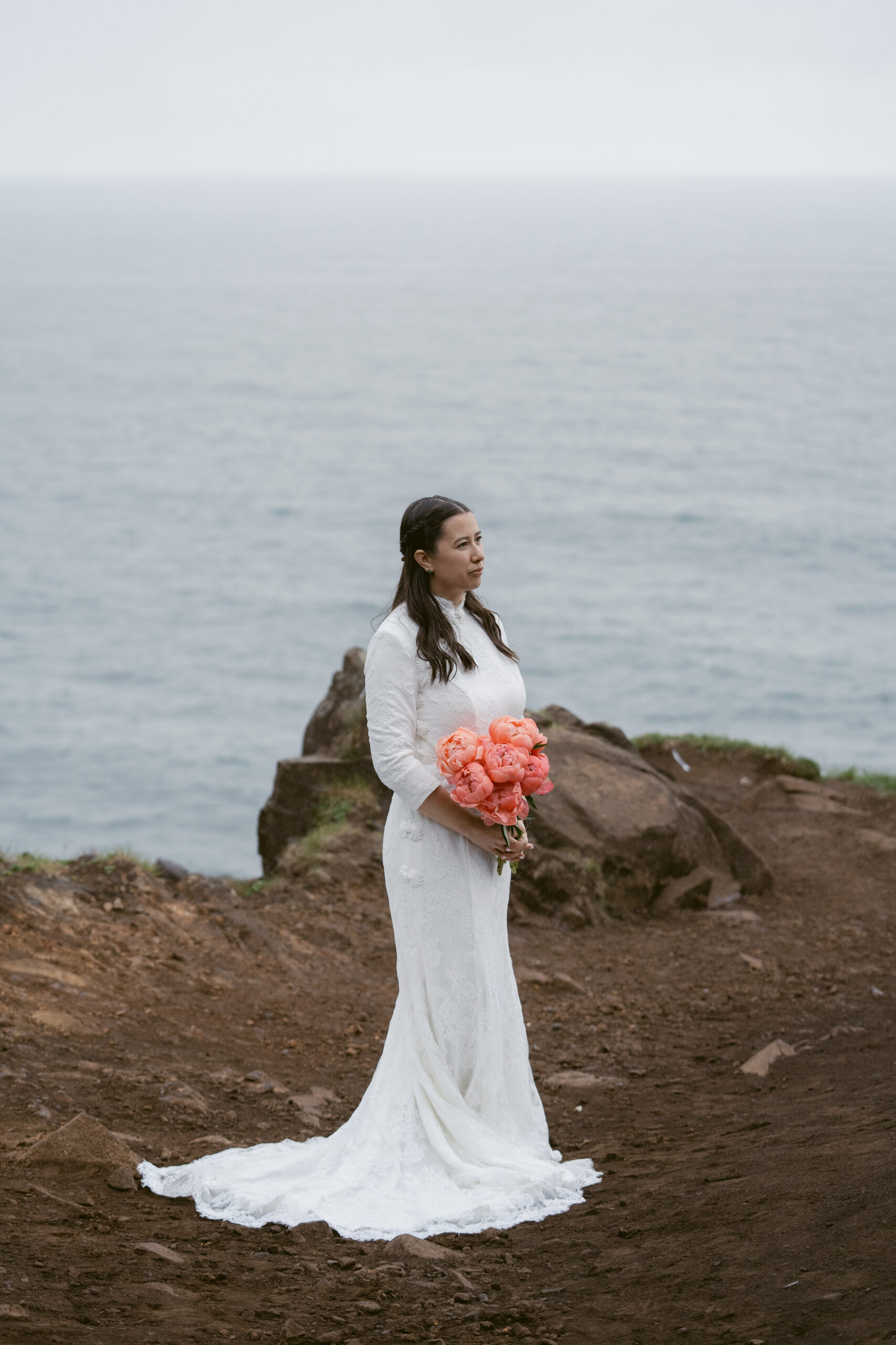 A bride stands on a rocky cliffside with the ocean behind her, holding a bouquet of coral peonies in her lace wedding dress.