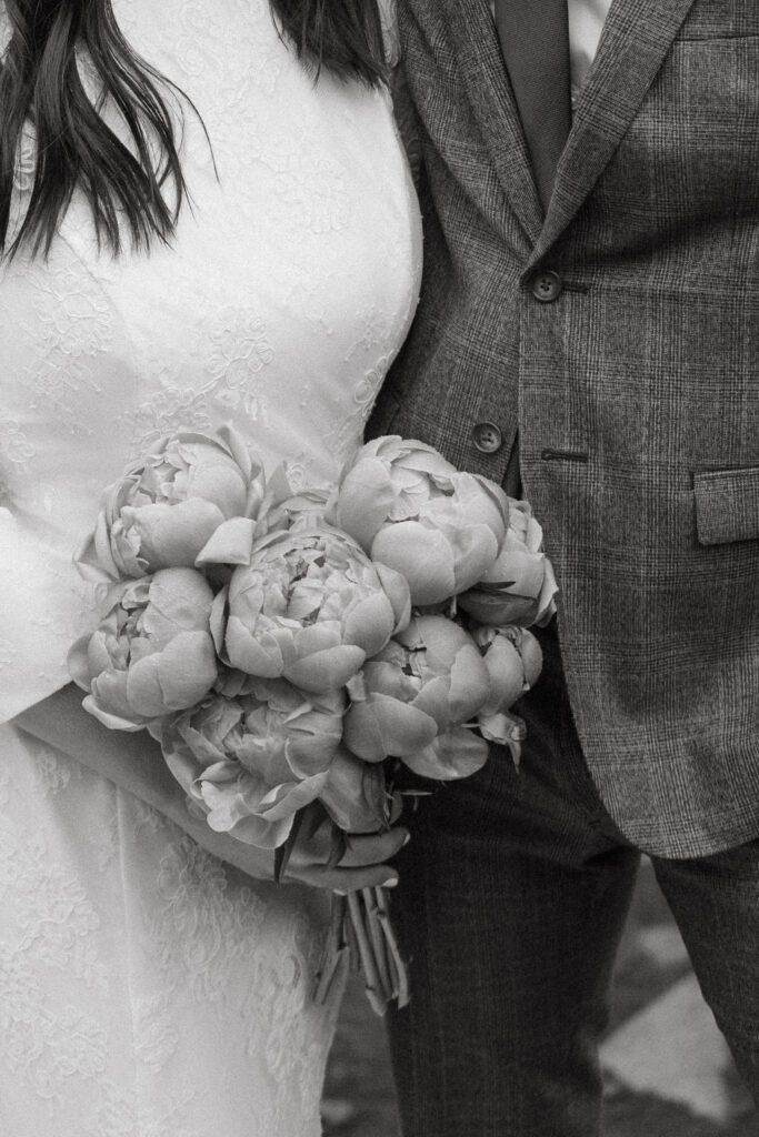 A close-up black-and-white image of a bride holding a bouquet of peonies, standing next to the groom in a plaid suit.