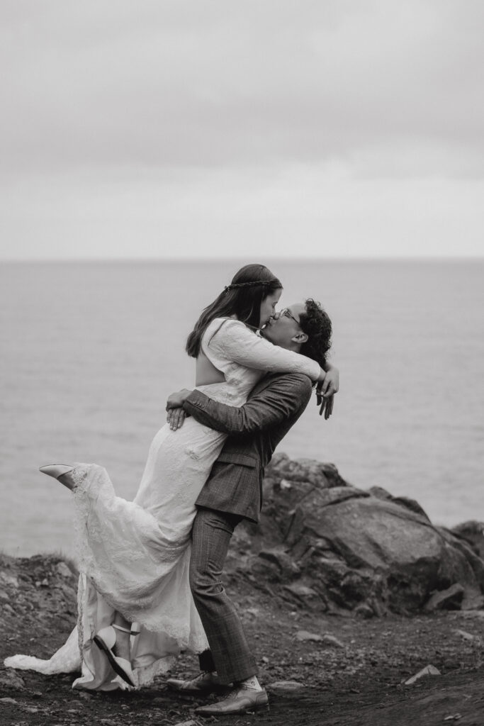 A joyful black-and-white moment of a groom lifting his bride as they kiss by the rocky coastline.