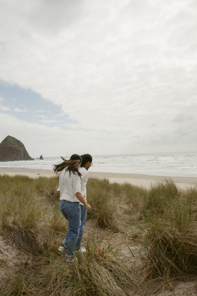 Couple walking through beach grass toward the ocean during their Oregon Coast elopement, wind in hair.