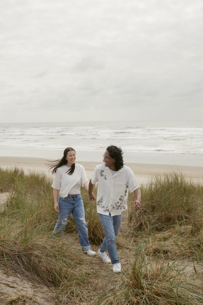 Couple holding hands and walking through beach grass during their Oregon Coast elopement, smiling at each other.