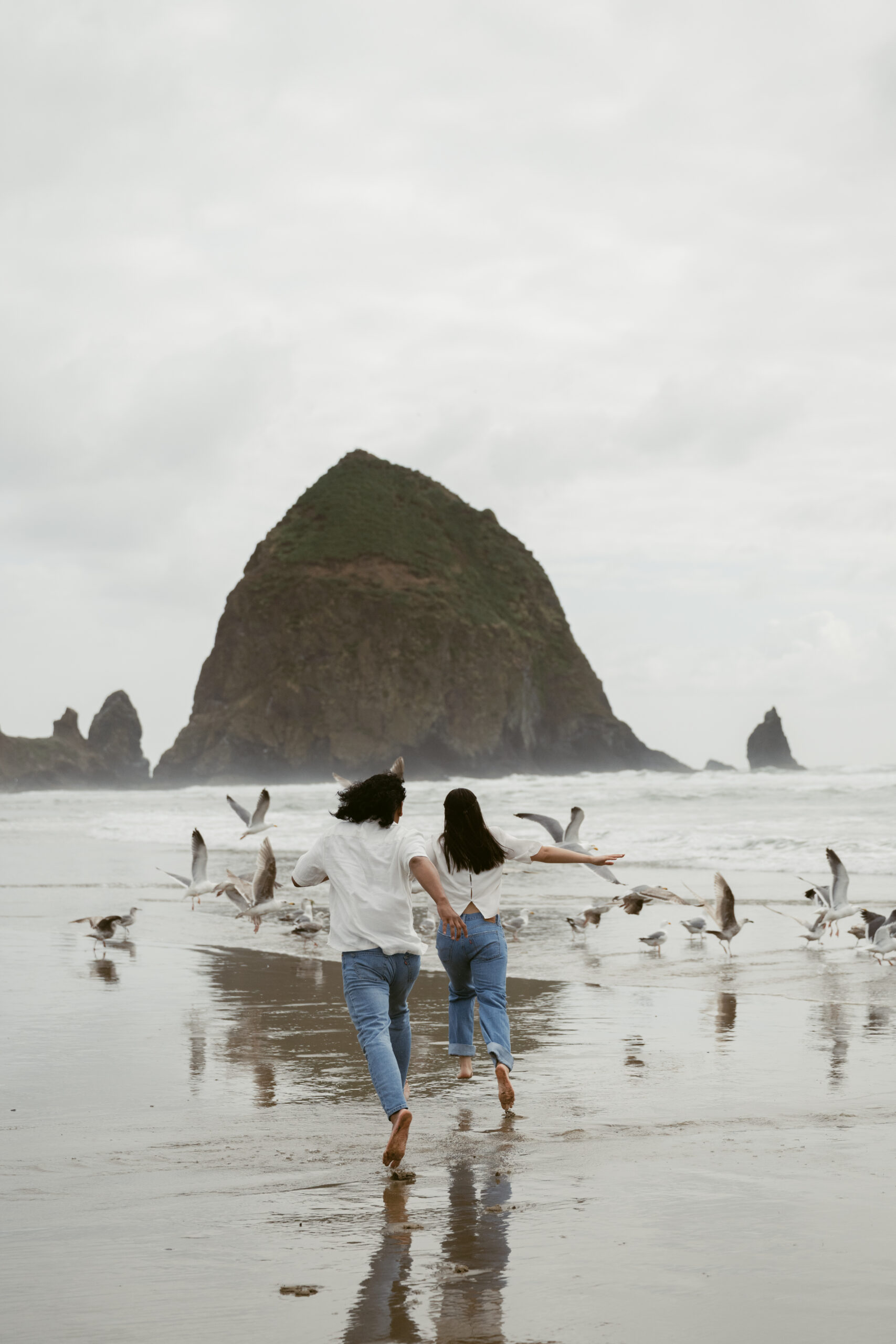 Couple running barefoot through seagulls during their Oregon Coast elopement, with Haystack Rock ahead.
